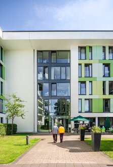 Moderne Wohnanlage mit grün-weißer Fassade und Glas-Eingang. Personen gehen auf den Eingang zu.