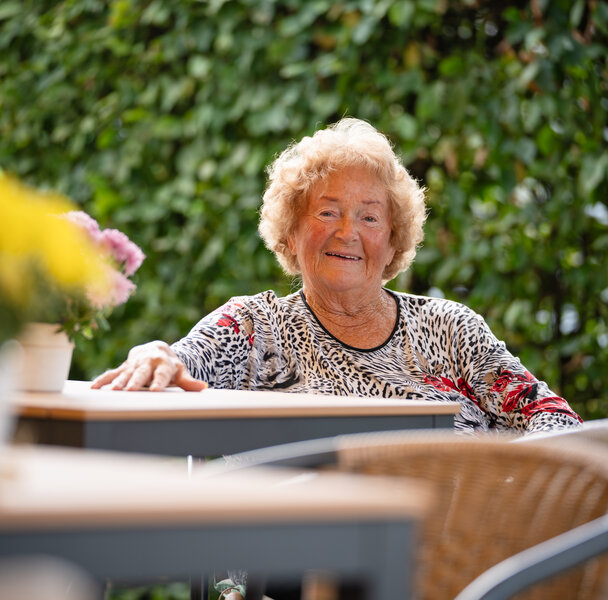 Lächelnde ältere Frau sitzt entspannt an einem Tisch im Freien.