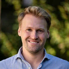 Lächelnder blonder Mann mit Bart und blau-weißem Hemd.