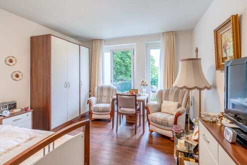 Helles möbliertes Zimmer in einer Wohnanlage mit Bett, Tisch und Sesseln.