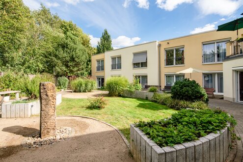 Sonnige Wohnanlage mit heller Fassade, grünem Garten, Kieswegen und Pflanzen.