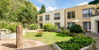 Sonnige Wohnanlage mit heller Fassade, grünem Garten, Kieswegen und Pflanzen.