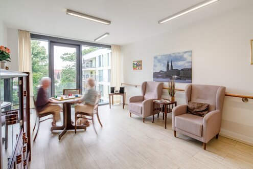 Zwei Senioren spielen im hellen Gemeinschaftsraum einer Wohnanlage ein Brettspiel am Fenster.