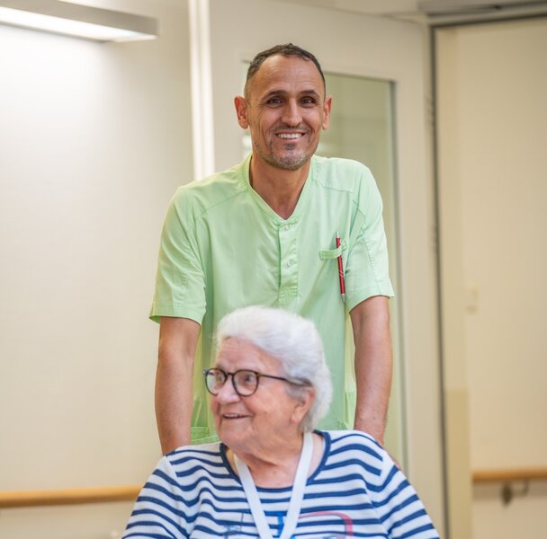 Pfleger schiebt lächelnde Seniorin im Rollstuhl in einer Wohnanlage.