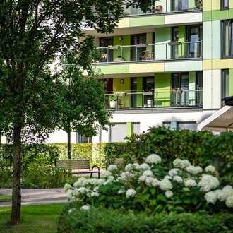 Grüne Wohnanlage mit Balkonen, umgeben von Bäumen und einer Parkbank.