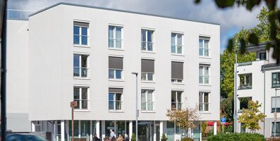 Außenansicht einer hellen Wohnanlage mit Senioren auf Bänken davor.