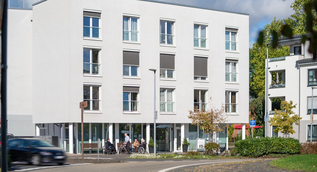 Außenansicht einer hellen Wohnanlage mit Senioren auf Bänken davor.