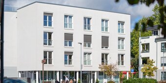 Außenansicht einer hellen Wohnanlage mit Senioren auf Bänken davor.