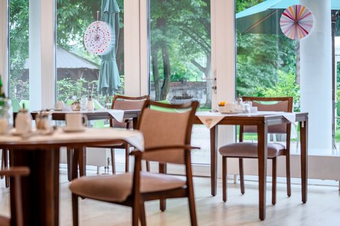 Speisesaal einer Wohnanlage mit gedeckten Tischen und großem Fensterblick in einen grünen Garten.