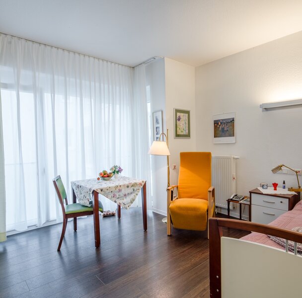 Wohnraum in einer Einrichtung: Bett, Esstisch, Sessel, viel Licht.