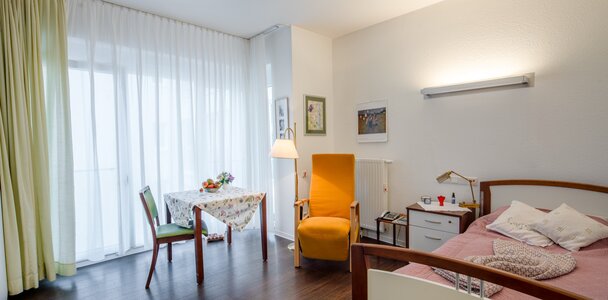 Wohnraum in einer Einrichtung: Bett, Esstisch, Sessel, viel Licht.