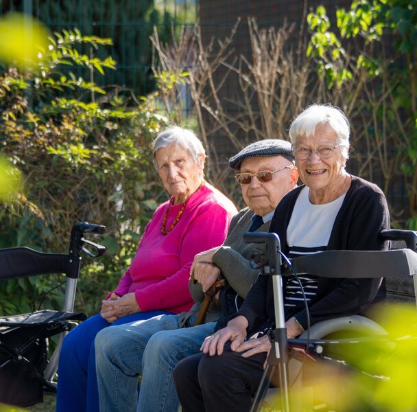 Drei ältere Bewohner einer Wohnanlage sitzen im Garten mit Rollatoren.
