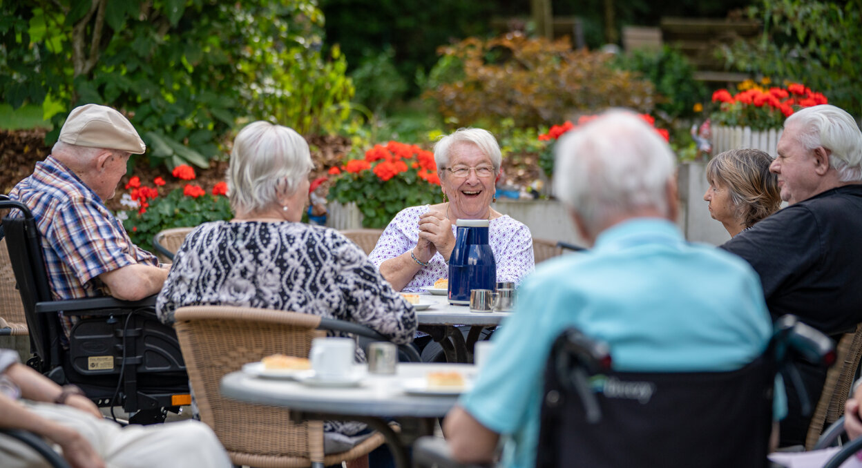 Lachende Senioren sitzen bei Kaffee und Kuchen im grünen Garten einer Wohnanlage.