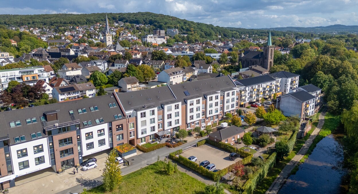 Moderne betreute Wohnanlage am Fluss, umgeben von einer Stadt mit Kirche und grünen Hügeln.