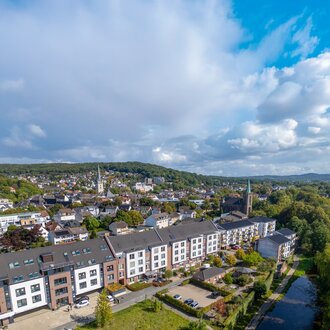 Moderne betreute Wohnanlage am Fluss, umgeben von einer Stadt mit Kirche und grünen Hügeln.
