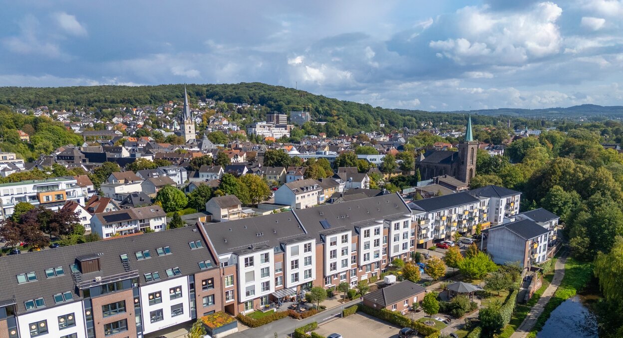 Moderne betreute Wohnanlage am Fluss, umgeben von einer Stadt mit Kirche und grünen Hügeln.
