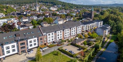 Moderne betreute Wohnanlage am Fluss, umgeben von einer Stadt mit Kirche und grünen Hügeln.