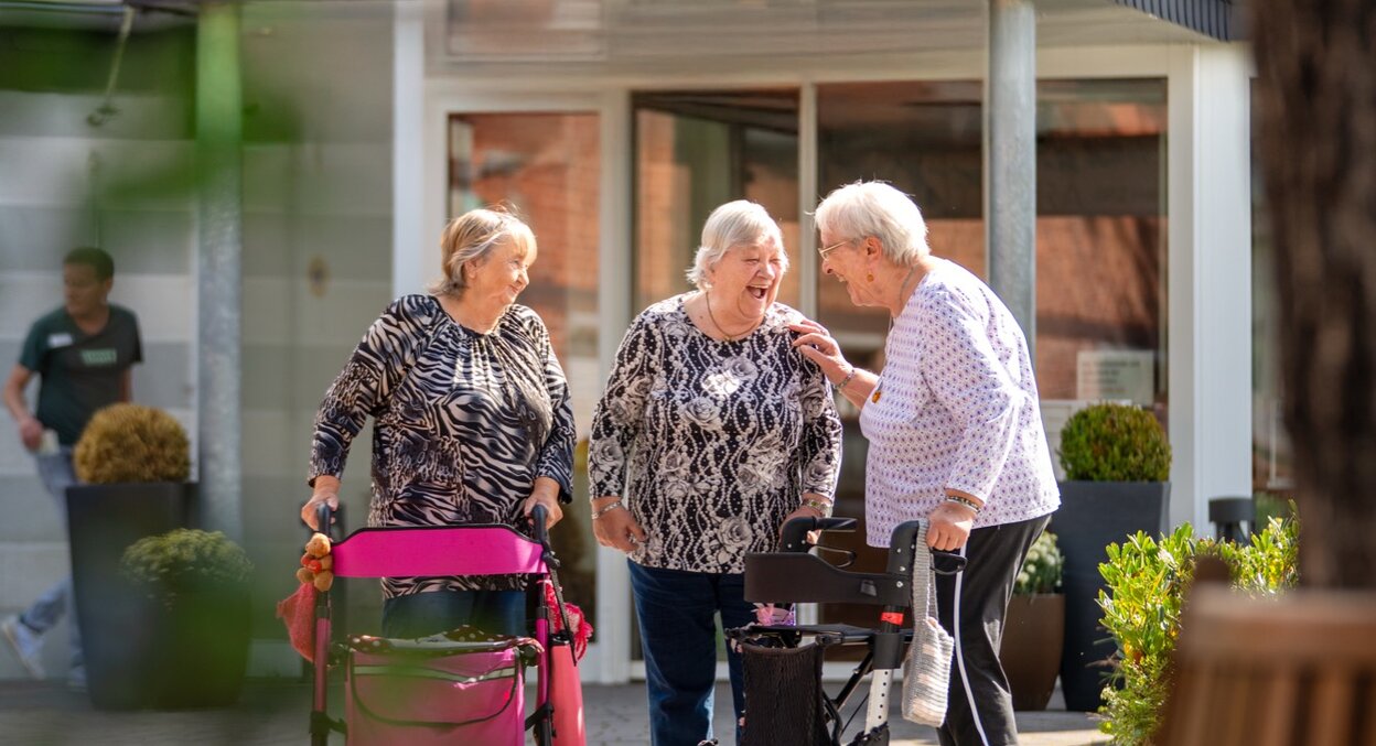 Drei lachende Seniorinnen mit Rollatoren vor einer Wohnanlage.