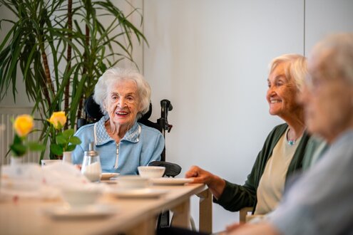 Drei lächelnde Seniorinnen sitzen zusammen an einem Tisch in einer Wohnanlage.