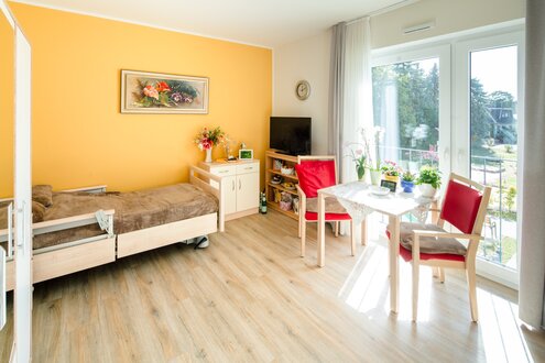 Helles Zimmer mit Pflegebett, Esstisch und großen Fenstern in einer betreuten Wohnanlage.
