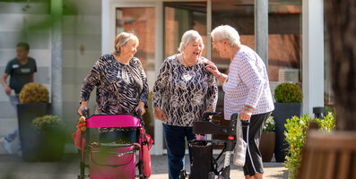 Drei lachende Seniorinnen mit Rollatoren unterhalten sich vor der Wohnanlage.
