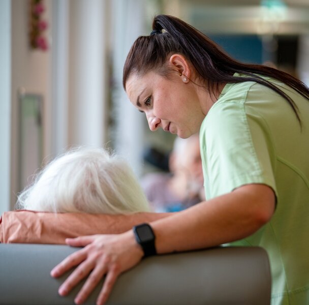 Pflegekraft beugt sich fürsorglich zu einer älteren Person in einer Wohnanlage.