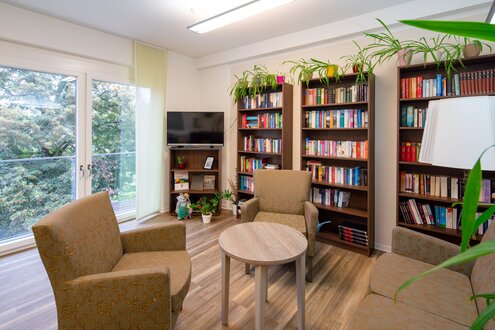 Gemütlicher Gemeinschaftsraum mit Bücherregalen, Sesseln und Fensterblick ins Grüne in einer Wohnanlage.