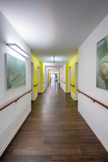 Langer, heller Flur in einer betreuten Wohnanlage mit Handläufen und gelben Akzenten.