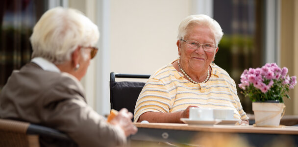 Zwei Seniorinnen sitzen lächelnd beim Kaffee draußen in einer Wohnanlage.