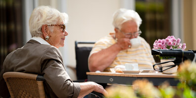 Zwei Senioren genießen Kaffee und Gebäck auf einer sonnigen Terrasse.