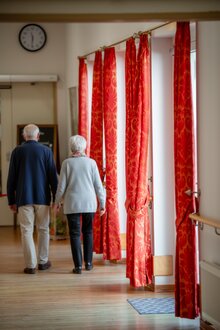Zwei ältere Menschen gehen durch den hellen Flur einer Wohnanlage mit roten Vorhängen.