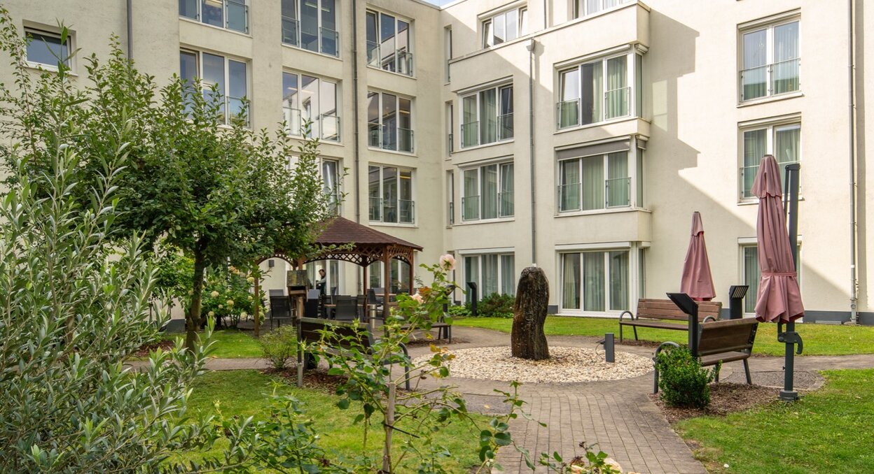 Innenhof einer Senioren-Wohnanlage mit grünem Garten, Wegen, Bänken und Pavillon.
