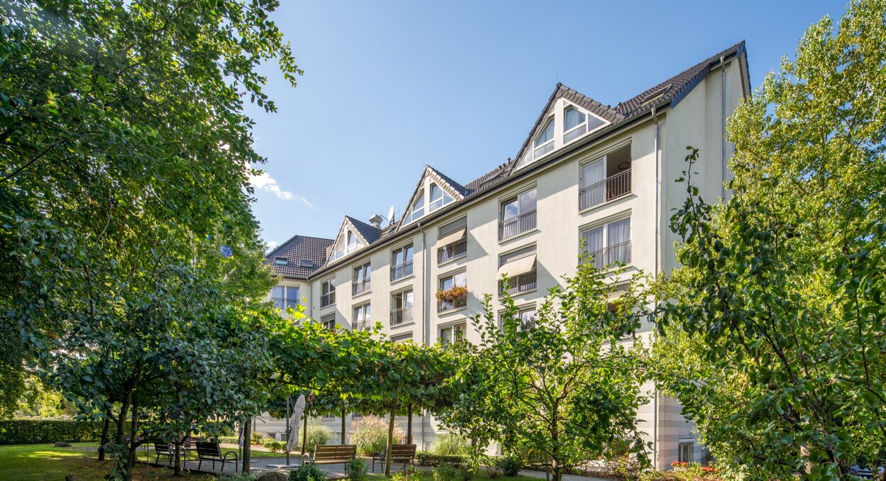 Mehrstöckiges Wohngebäude mit Balkonen, umgeben von gepflegtem Grün und Sitzbänken.