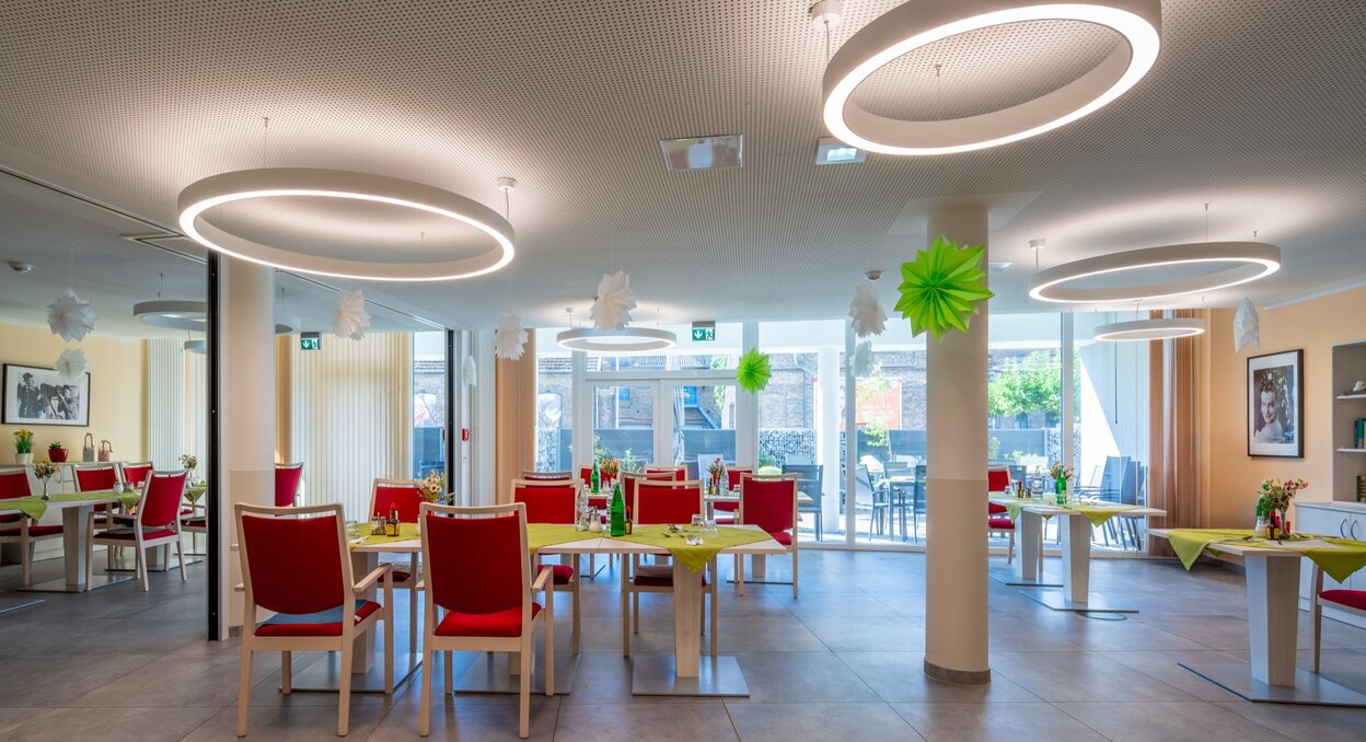 Heller Speisesaal mit Tischen, roten Stühlen und großen Fenstern in einer Wohnanlage.