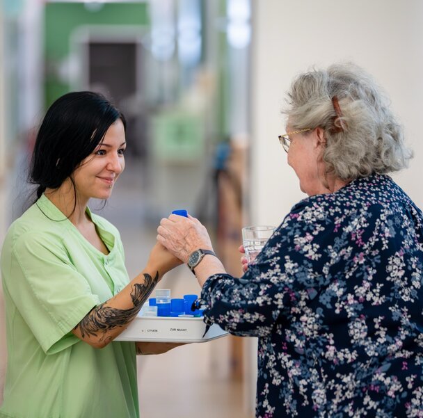 Pflegekraft reicht Seniorin in betreuter Wohnanlage Medikamente mit Wasser.