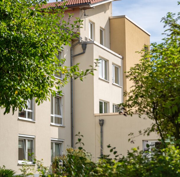 Helles Gebäude für betreutes Wohnen mit rotem Dach, von grünen Bäumen umrahmt.