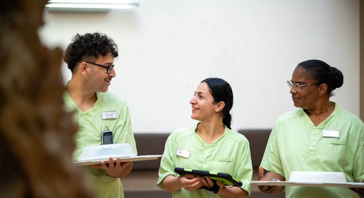 Drei lächelnde Mitarbeiter in grünen Uniformen mit Tablets und Essenstrays im Gespräch.