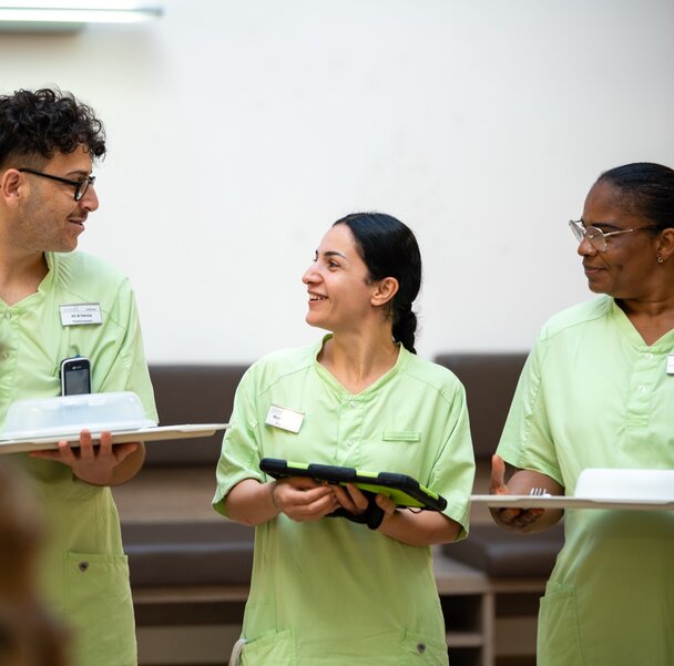 Drei lächelnde Mitarbeiter in grünen Uniformen mit Tablets und Essenstrays im Gespräch.