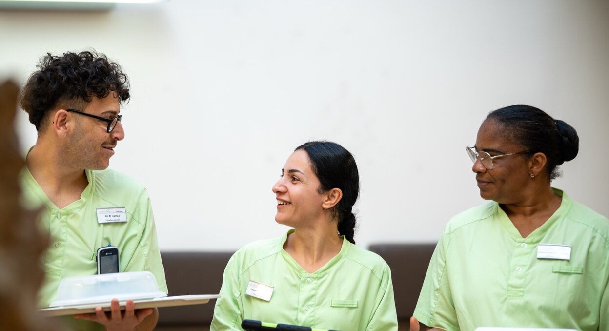 Drei lächelnde Mitarbeiter in grünen Uniformen mit Tablets und Essenstrays im Gespräch.