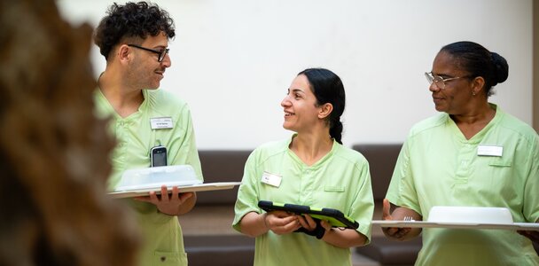 Drei lächelnde Mitarbeiter in grünen Uniformen mit Tablets und Essenstrays im Gespräch.
