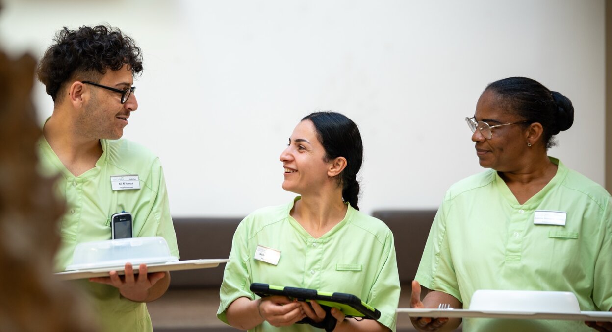 Drei lächelnde Mitarbeiter in grünen Uniformen mit Tablets und Essenstrays im Gespräch.