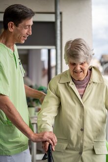 Mitarbeiter begleitet Seniorin mit Gehhilfe in Wohnanlage.