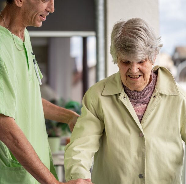Mitarbeiter begleitet Seniorin mit Gehhilfe in Wohnanlage.