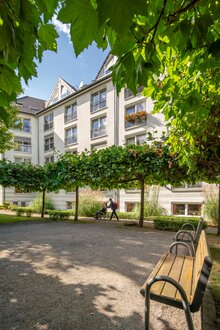 Betreutes Wohnen: Moderne Wohnanlage mit Grünanlage. Eine Person schiebt einen Rollstuhl.