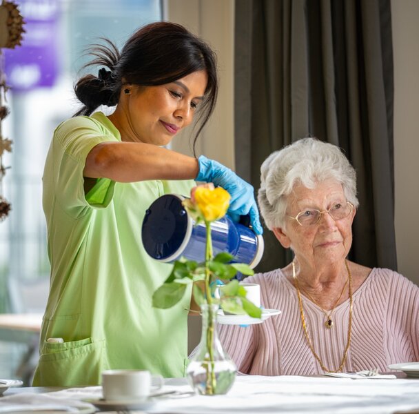 Pflegekraft gießt Seniorin in einer Wohnanlage ein Getränk ein.