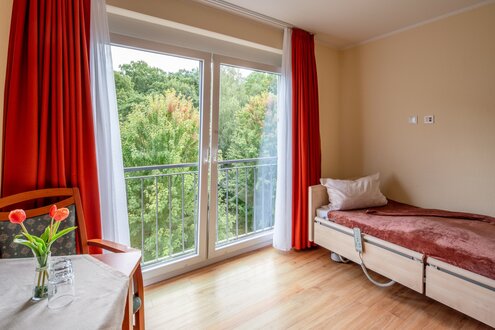 Helles Zimmer in betreutem Wohnen mit Pflegebett, Tisch und Balkontür zu grünen Bäumen.