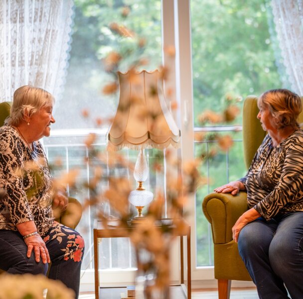 Lachende Seniorinnen unterhalten sich in bequemen Sesseln einer Einrichtung.