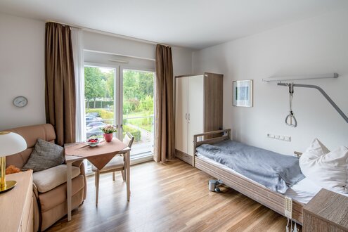 Helles Bewohnerzimmer einer Pflegeeinrichtung mit Pflegebett, Sessel und Esstisch am Fenster.