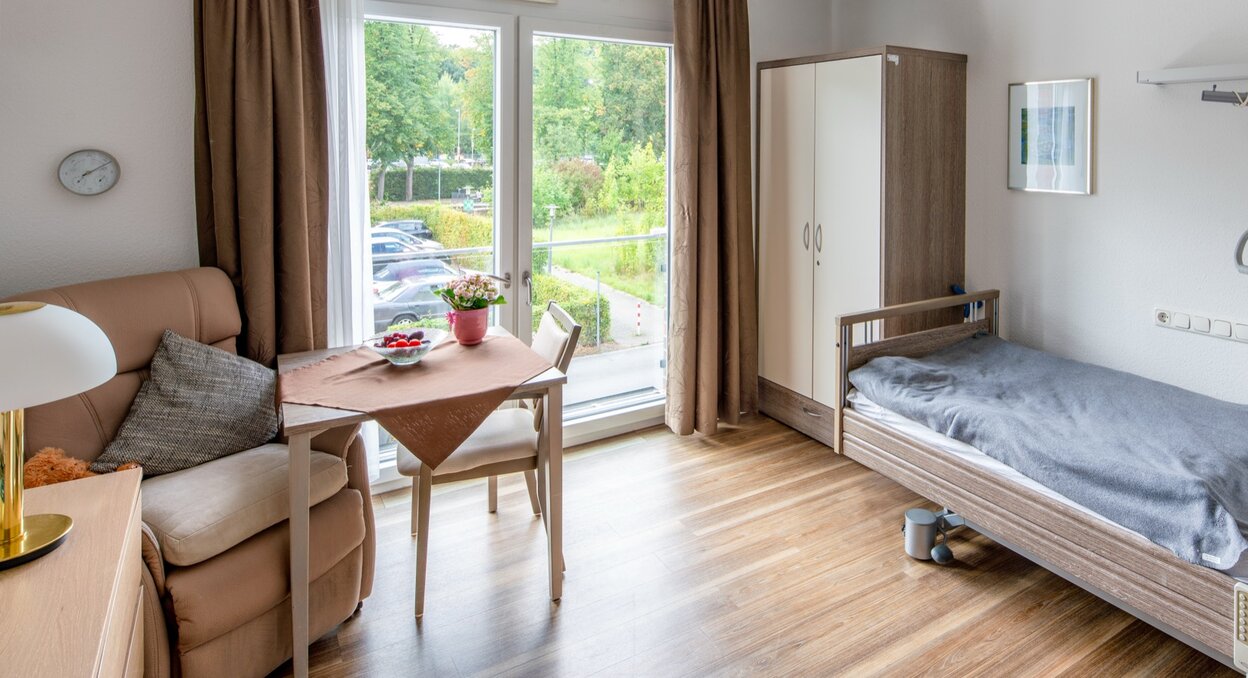 Helles Bewohnerzimmer einer Pflegeeinrichtung mit Pflegebett, Sessel und Esstisch am Fenster.