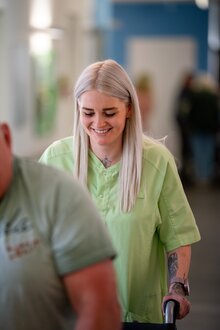 Lächelnde Betreuungskraft in grüner Uniform schiebt Gehhilfe.
